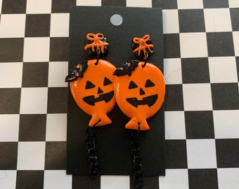 May include: A pair of orange and black Halloween-themed earrings. Each earring features a jack-o'-lantern balloon with a black bat and a black bow on top. The earrings are attached to black chains.