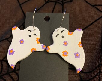 May include: A pair of white ghost-shaped earrings with purple and orange flower patterns. The earrings are made of polymer clay and have silver hoop hooks.