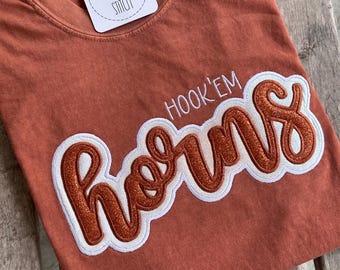 May include: A rust-colored short-sleeve t-shirt featuring the embroidered text HOOK'EM HORNS in a white and glittery copper script font. A white square tag with the text THE JOLLY STITCH is attached to the inner neckline of the garment.