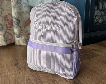 May include: A small, seersucker backpack in lavender and white stripes. The word "Sophia" is embroidered in white script on the front. Features a lavender zipper and mesh side pocket. The backpack is sitting on a wooden floor.