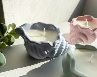 May include: Three decorative candles in the shape of seashells. The candles are in shades of light blue, pink, and green. The candles are sitting on a white surface.