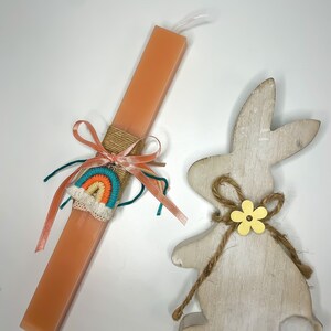 Può includere: Una candela pasquale color pesca con una decorazione in filato arcobaleno e un fiocco marrone. Una statuetta di coniglio in legno bianco con un fiore giallo e spago marrone.