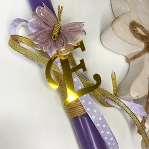 Può includere: Una candela viola decorata con un'iniziale dorata, una farfalla e nastri. La farfalla è viola chiaro con dettagli dorati. La candela è avvolta con spago e adornata con nastri dorati e bianchi a pois.