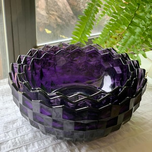 May include: A deep purple glass bowl with a geometric, faceted design. The bowl has a wide, open shape and sits on a white surface. The bowl's edges have a dark, almost black, trim.