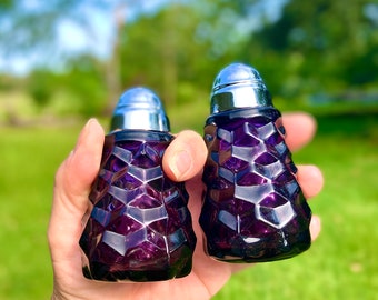 May include: A pair of vintage purple glass salt and pepper shakers with a geometric design. Each shaker has a silver metal lid. The shakers are held in a hand against a blurred green and blue background.