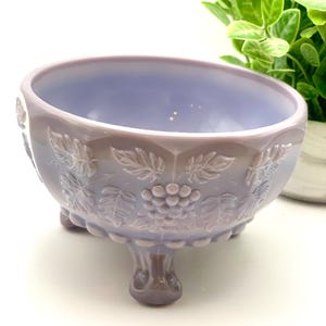 May include: A lavender-colored, footed bowl with a grape and leaf design. The bowl has a scalloped rim and three decorative feet. The grape and leaf pattern is raised and adds texture to the bowl's surface. The bowl is likely made of glass.