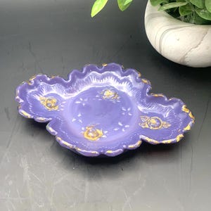 May include: A small, purple ceramic dish with a scalloped edge and gold trim. The dish features gold floral designs and is likely used for holding small items. The background is a dark surface with a decorative plant.