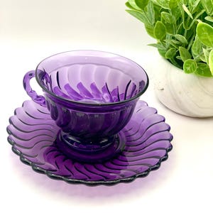 May include: A purple glass teacup and saucer set. The cup and saucer have a swirled design. The cup has a handle and sits on a pedestal base. The saucer has a scalloped edge. A small green plant in a white pot is in the background.