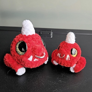 May include: Two plush red dragon toys with white horns, white claws, and large black and gold eyes. Each dragon has a silver nose ring. The toys are handmade and have a soft, textured appearance.