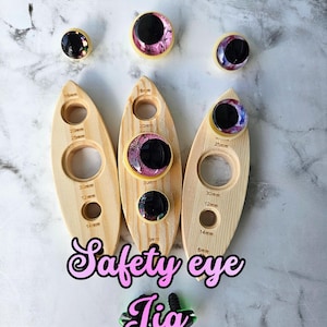May include: A selection of wooden safety eye jigs with different hole sizes, and several colourful safety eyes. The text "Safety eye Jig" is displayed in a pink font. The jigs have measurements in millimetres.