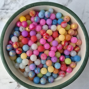 Op de afbeelding: Een ronde kom met een donkergroene rand gevuld met een grote verzameling kleine, bolvormige kralen in diverse kleuren, waaronder roze, blauw, geel, paars en wit. Veel van de kralen hebben een subtiele glitterende of gemarmerde afwerking.