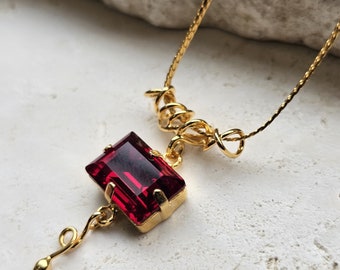 May include: A gold necklace with a red gemstone pendant. The pendant is a rectangular shape with a gold setting. The necklace has a delicate chain with a twisted design.