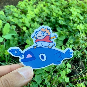 May include: A blue whale sticker with a small, red-shirted character riding on top. The character has blonde hair and large, round eyes.