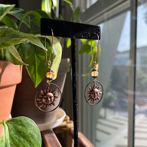 May include: A pair of gold-toned earrings with a sun face charm and a yellow bead. The earrings are hanging from a black display stand.