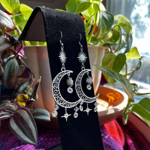 May include: Silver crescent moon earrings with dangling stars and crystals. The earrings feature a white moonstone bead on each earring.