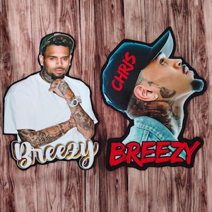 May include: Two die-cut images of a man with tattoos, one in a white shirt and the other in a denim jacket and a black cap with the word "CHRIS". The word "Breezy" is written in a stylized font on both images. The background is a wood grain pattern.