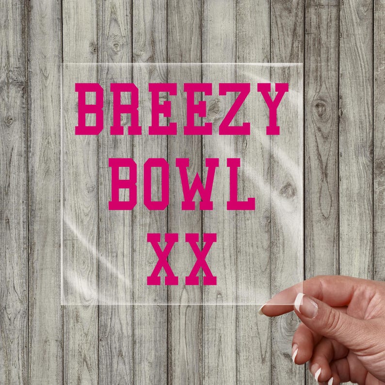 May include: A clear, square decal with the words "BREEZY BOWL XX" in bright pink, block letters. The decal is held up by a hand against a wooden background.
