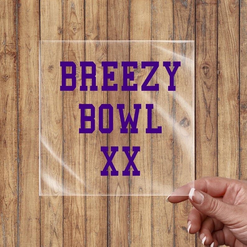 May include: A clear, square acrylic sign with the words "BREEZY BOWL XX" in purple block letters. The sign is held by a hand with manicured nails against a wooden background.