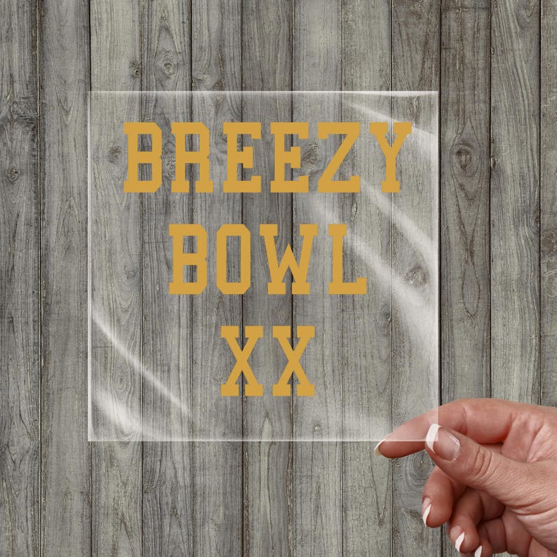 May include: A clear, square acrylic sign with the words "BREEZY BOWL XX" in gold lettering. The sign is held by a hand with manicured nails against a wooden background.