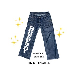 May include: Blue denim jeans with white block letters spelling "BREENZY" down the left pant leg. The jeans are 16 x 3 inches. The jeans have pockets and a button closure.