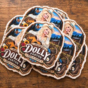 May include: A stack of die-cut stickers featuring a portrait of Dolly Parton wearing a cowboy hat, playing an acoustic guitar, and surrounded by sunflowers and butterflies. The text reads Dolly Parton Queen of Country in a decorative banner.
