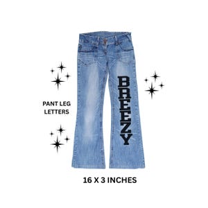 May include: Light blue denim jeans with a flared leg design. The word "BREEZY" is printed vertically in black on the right pant leg. The jeans measure 16 x 3 inches.