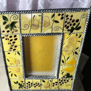 May include: A decorative photo frame with a yellow tile design featuring fruit and leaf motifs. The frame has a silver rope-like border and a square opening for a photo. The frame is approximately 8 inches tall and 6 inches wide.