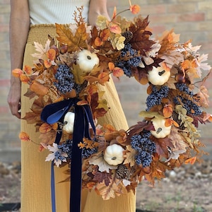 May include: A circular autumn wreath featuring artificial maple leaves in shades of orange, brown, and yellow, accented with small white pumpkins, clusters of dark blue berries, and pinecones. A long, dark navy blue velvet ribbon hangs from the side.