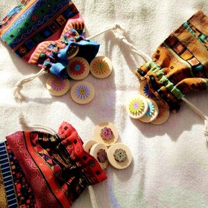 Può includere: Tre sacchetti a cordoncino colorati con gettoni di legno a forma di fiore all'interno. I sacchetti hanno un motivo geometrico con colori rosso, blu, giallo e verde. I gettoni hanno una varietà di colori e disegni.