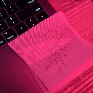 May include: A pink fabric patch with a design of small, round, silver-coloured rhinestones arranged in the shape of a palm tree. The patch is lying on a pink surface.