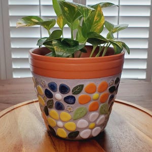 May include: A decorative ceramic flower pot with a mosaic design. The pot features a terracotta rim and is filled with a green leafy plant. The mosaic design includes colorful, rounded tiles in shades of yellow, blue, orange, and green.