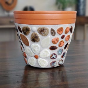 May include: A terracotta flower pot decorated with a mosaic of white, brown, and orange tiles. The tiles are arranged in a floral pattern.