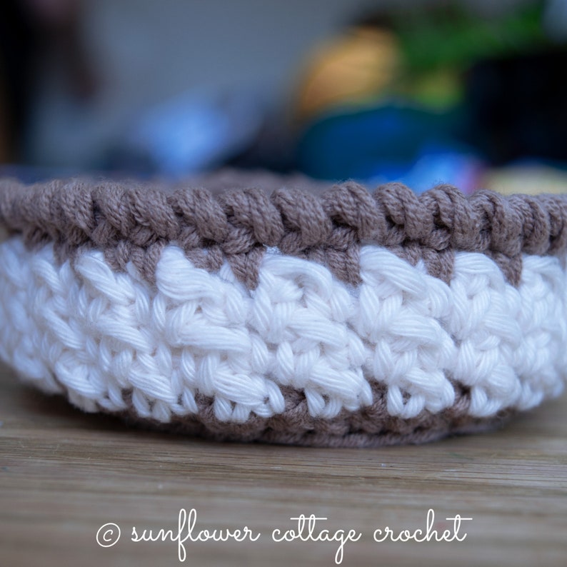 May include: A crocheted basket with a brown and white striped pattern. The basket is made with a thick yarn and has a rounded shape.