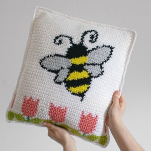 Puede incluir: Un coj&iacute;n cuadrado de crochet blanco con un gran dise&ntilde;o de abeja en negro, amarillo y gris. El borde inferior tiene una fila de tulipanes rosas con tallos y hojas verdes. El coj&iacute;n est&aacute; sostenido por manos.