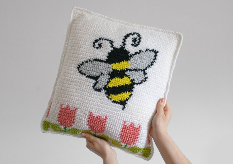 Puede incluir: Un coj&iacute;n cuadrado de crochet blanco con un gran dise&ntilde;o de abeja en negro, amarillo y gris. El borde inferior tiene una fila de tulipanes rosas con tallos y hojas verdes. El coj&iacute;n est&aacute; sostenido por manos.