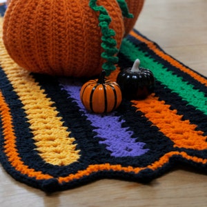 May include: A crocheted rug with black, orange, yellow, green, and purple stripes. There are three small crocheted pumpkins on the rug, two orange and one black.
