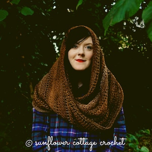 May include: A woman wearing a brown crocheted cowl scarf with a plaid shirt underneath. The scarf is draped around her neck and shoulders. The woman is standing in front of a dark green background.