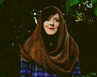 May include: A woman wearing a brown crocheted cowl scarf with a plaid shirt underneath. The scarf is draped around her neck and shoulders. The woman is standing in front of a dark green background.