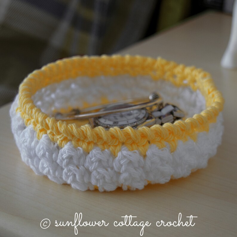 May include: A crocheted bowl with a white and yellow striped design. The bowl is filled with a watch and a silver pin.