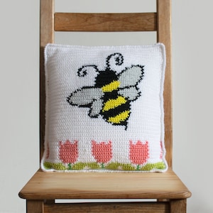 Puede incluir: Coj&iacute;n blanco de ganchillo con un gran dise&ntilde;o de abeja en negro, amarillo y gris. El coj&iacute;n presenta una fila de tulipanes rosas con tallos y hojas verdes a lo largo del borde inferior. El coj&iacute;n est&aacute; colocado en una silla de madera.
