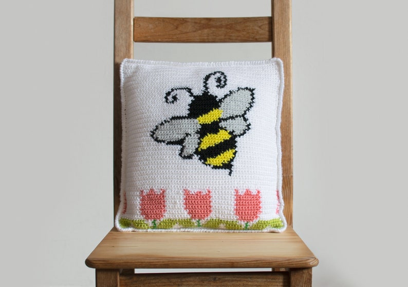 Puede incluir: Coj&iacute;n blanco de ganchillo con un gran dise&ntilde;o de abeja en negro, amarillo y gris. El coj&iacute;n presenta una fila de tulipanes rosas con tallos y hojas verdes a lo largo del borde inferior. El coj&iacute;n est&aacute; colocado en una silla de madera.