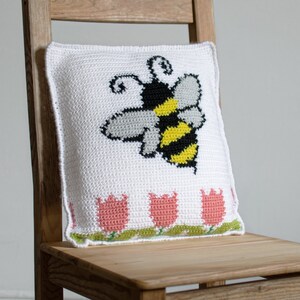 Puede incluir: Coj&iacute;n blanco de ganchillo con un gran dise&ntilde;o de abeja en negro, amarillo y gris. El coj&iacute;n presenta una fila de tulipanes rosas con hojas verdes a lo largo del borde inferior. Est&aacute; colocado sobre una silla de madera.