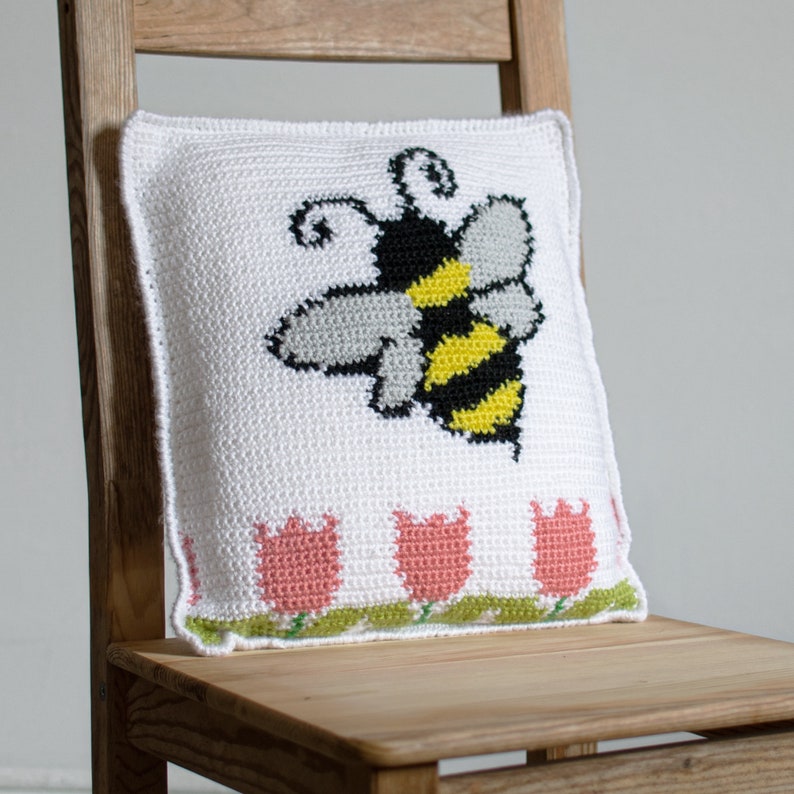 Puede incluir: Coj&iacute;n blanco de ganchillo con un gran dise&ntilde;o de abeja en negro, amarillo y gris. El coj&iacute;n presenta una fila de tulipanes rosas con hojas verdes a lo largo del borde inferior. Est&aacute; colocado sobre una silla de madera.