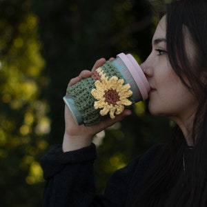 May include: A green crochet coffee cup cozy with a yellow and brown sunflower design. The cozy is wrapped around a light blue and pink travel mug. The image is set against a blurred green background.