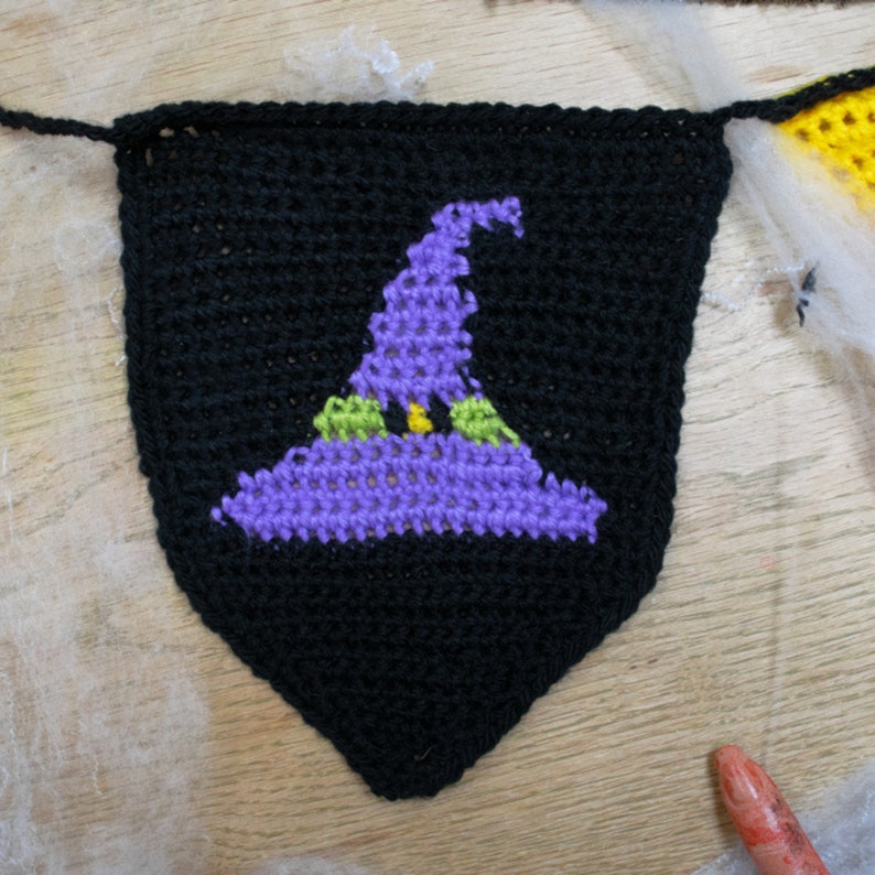 May include: A black crocheted witch hat banner with a purple hat and green accents. The hat is on a black triangle banner.