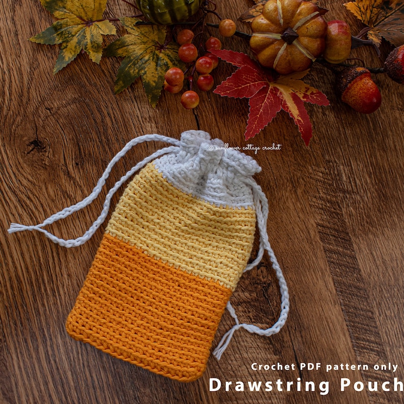 May include: Crocheted drawstring pouch in the shape of a candy corn. The pouch is white, yellow, and orange. The pouch is lying on a wooden surface with fall leaves and a small pumpkin.