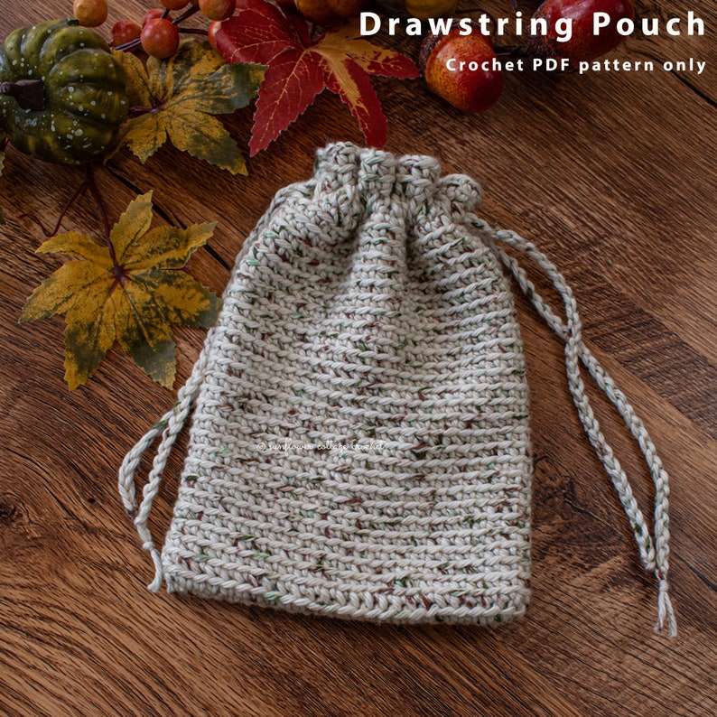 May include: A beige crocheted drawstring pouch with a brown and green speckled pattern. The pouch is on a wooden surface.