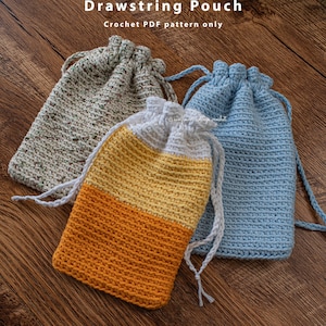 Puede incluir: Tres bolsas de cordón de crochet en diferentes colores. Las bolsas están hechas con un punto sencillo y tienen un cordón de ajuste blanco. Las bolsas están sobre una superficie de madera.