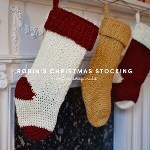 Op de afbeelding: Drie gehaakte kerstsokken hangen aan een witte schouw. De sokken zijn rood, wit en goud. De tekst "ROBIN'S CHRISTMAS STOCKING" is zichtbaar op de schouw.