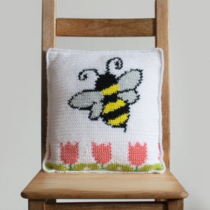 Puede incluir: Un coj&iacute;n cuadrado blanco con un dise&ntilde;o de abeja de ganchillo en negro, amarillo y gris. El coj&iacute;n tiene un borde de tulipanes rosas con tallos y hojas verdes. El coj&iacute;n est&aacute; colocado sobre una silla de madera.